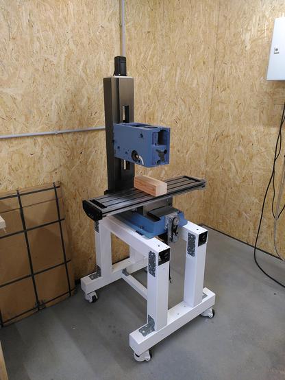 Blue milling machine with a white stand sitting in the corner of a work shop. Walls are tan OSB (wood) panels and the floor is grey concrete.