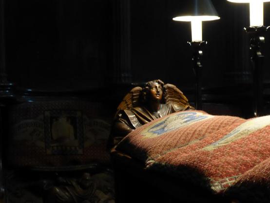 Carved wooden angel looks up towards quire lights. Black background.