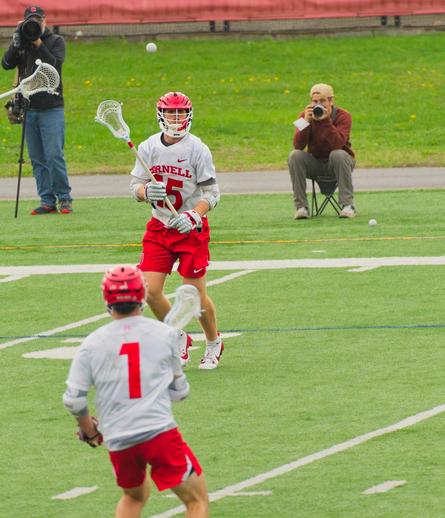 On the 1/3 line from the left two men are lined up,  both wearing red helmets, red shorts, white jerseys with red numbers and lacrosse sticks.  The ball is hanging in the area between them.  Behind them count two photographers taking pictures from the alternate sidelines and numerous yellow dandilions sprouting in the margin