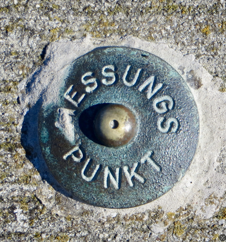 Ein Vermessungspunkt aus Messing, er ist kreisund und es steht in Großbuchstaben "Messungspunkt" rund um die Halbkugel in der Mitte. Das M ist von ganz feinem hellen Sand bedeckt.