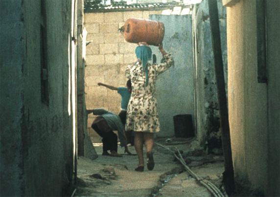 Filmstil aus Palestinian Women, ©Association Jocelyne Saab, Marseille. Eine Frau trägt eine Gasflasche auf dem Kopf durch eine Enge Gasse. Sie steht mit dem Rücken zur Kamera und läuft in die Richung von zwei spielenden Kinder.