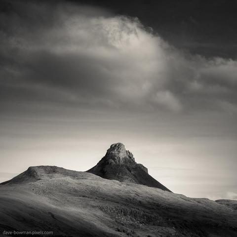 In this monochrome photograph of the Scottish Highlands, a serene scene unfolds as clouds gently gather above the iconic Stac Pollaidh. The minimalist style captures the essence of the landscape, showcasing the warm-toned tip of the mountain rising proudly against the sky. As the cloud begins to form above its peak, a sense of tranquility and grandeur envelops the frame, inviting contemplation and appreciation for the majestic beauty of nature.