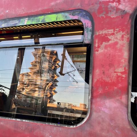 Ein S-Bahn-Fenster umrahmt von rostig-abblätternder roter Farbe, in dem sich der vom Sonnenuntergang orange gefärbte Kaktusturm - ein stachelig aussehendes Hochhaus - verzerrt spiegelt.