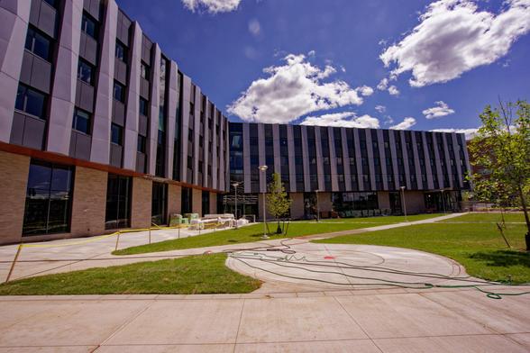 A building that would be L-shaped except the angle is more than 90 degrees,  maybe more like 135 degrees,  and there's a plaza of mostly gree grass with a few trees except for some concrete sidewalks and a concrete round area.