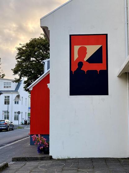 A street scene showing a bit of a red house and a white wall. The wall has what looks to be movie art poster painted on it. The “poster” has a silhouette looking at a projection of itself. The silhouette looks a bit like Michael Caine. 