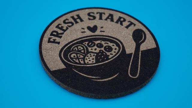 Korkuntersetzer lasegraviert mit der Aufschrift Fresh Start und einer Müslischüssel mit einem kleinen Herz drüber und einem Löffel daneben.