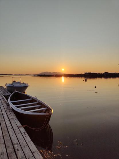 Photo de coucher de soleil. Il était  plus de 23h en Norvège en juillet. Paysage de fjord. Au premier plan, une barque et un bateau. Le soleil est en train de se coucher. Il se reflète dans l'eau, au ras de l'horizon, diffusant une douce couleur orangé. Le reste du ciel est d'un bleu très pale, sans nuage. L'eau est presque immobile. Tout cela donne une ambiance de grande sérénité.