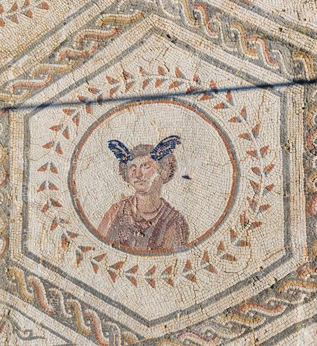 Mosaic of Hermes inside a circle decorated with an outer ring of leaf-shapes. He is wearing a chlamys cloak pinned over his right shoulder. His hair is short, about ear-length, and he has blue wings growing from his head.