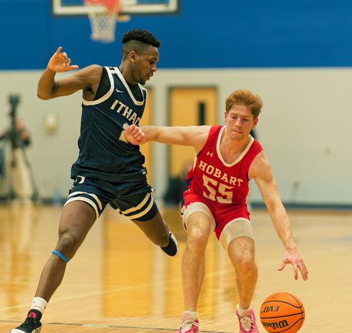 A white man with a red Jersey that says Hobart 55 is dribbling the basketball,  at the moment right near the floor and has his right arm (your left) extended to keep away a black man in a blue jersey that says Ithaca 2.  At the upper left the basket and a slice of the backboard is visible and a blurred out camera on a tall mount as well as some doors -- the back wall is light grey on the bottom and dark blue above.