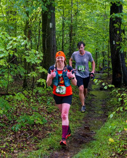A woman with braided twintails and a red jersey and blue vest makes a double thumbs up while a man in a grey shirt behind her is looking fierce as they run past two trees with square rectangular blazes.