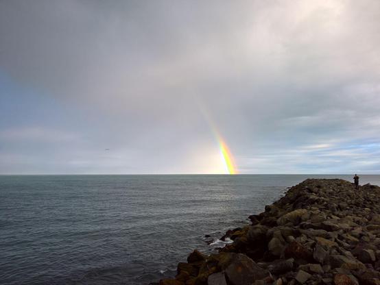 An einem Steindeich mit Blick aufs Wasser, am Horizont ist ein Regenbogen, der im Meer verschwindet, zu sehen. Der Himmel ist bewölkt, das Wasser ist grau.