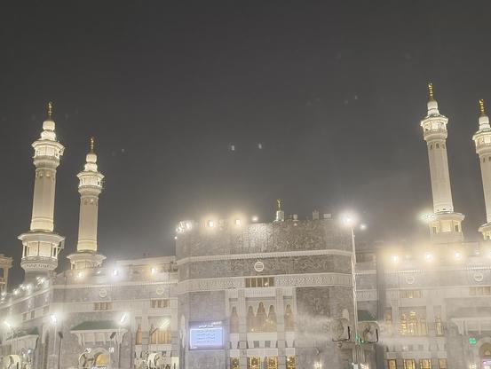 A wide-angle, low-light shot captures the exterior of the Masjid al-Haram (The Sacred Mosque) in Mecca, Saudi Arabia. The central section of the building is prominently featured, with its intricately decorated facade illuminated by numerous spotlights. The soft glow of the lights creates a hazy, ethereal effect in the humid night air. Two tall, white minarets with golden domes stand on either side of the building, reaching up into the dark, cloudy sky. Faint reflections of light are visible on the wet ground in the foreground.