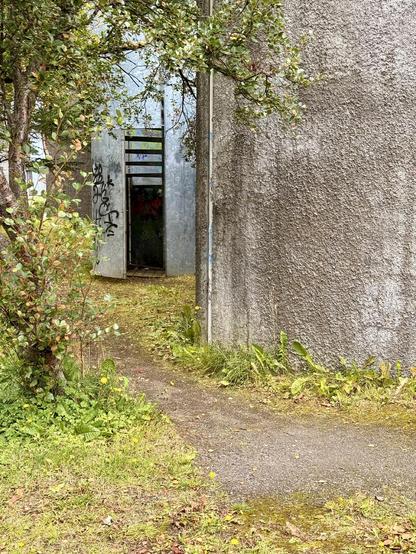 An entrance to a spiral staircase is seen on the side of a building next to a trail. It looks abandoned. 