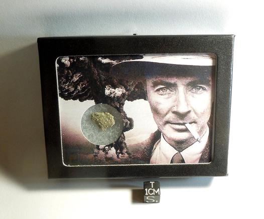 A display box. The display contains a small greenish piece of trinitite and a black and white collage photo of Oppenheimer and a mushroom cloud. 