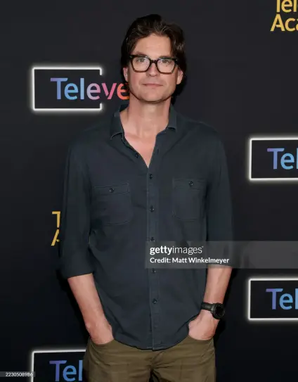 Gale Harold on the red carpet. He wears green pants, a black denim shirt and black framed glasses.