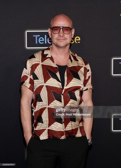 Peter Page on the red carpet. He wears plack pants, a red/black/beige patterned shirt and black framed red tinted sun glasses.