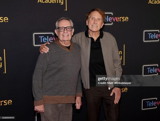 Daniel Lipman and Ron Cowen standing arm in arm next to each other on the red carpet. Daniel wears brown pants, a grey-brown jumper and black-framed glasses. Ron wears black pants, a black shirt and a black and white patterned jacket.