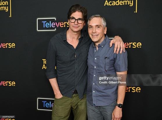 Gale Harold and Scott Lowell standing arm in arm next to each other on the red carpet. Gale wears green pants, a black denim shirt and black framed glasses. Scott wears blue jeans and a blue shirt with short sleeves.