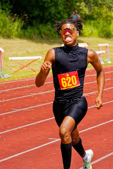 Black man running black shirt and black shorts and black socks running really hard with chroma sunglasses;  way in the background somebody is photographing this event from the other side.