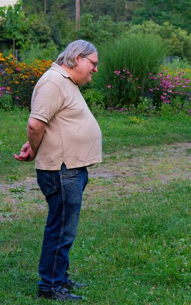 Man with grey hair and a beige shirt that bulges over his torso and his hands behind his back and blue jeans is seen in profile and behind him is a garden that includes an ornamental grass,  false sunflowers and coneflowers