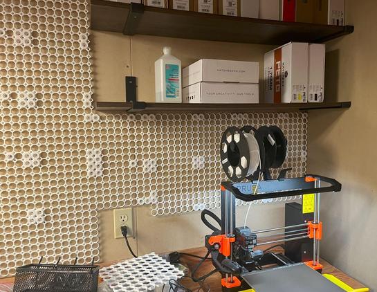 A basement corner, one wall covered in Multiboard storage tiles. In the top right, a conventional shelf, holding 3dprinting filament and a bottle of IPA (the cleaning kind). Bottom right, a pimped up Prusa i3 Mk3s, ready for duty.
