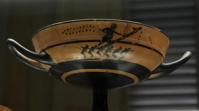 Phallophoria, carrying of a large phallus, depicted on an Attic black-figured kylix cup. Six presumably male figures carry a large bearded figure holding two grapevines and a man-sized phallus. The phallus has an eye on the glans and other decorations along the shaft.