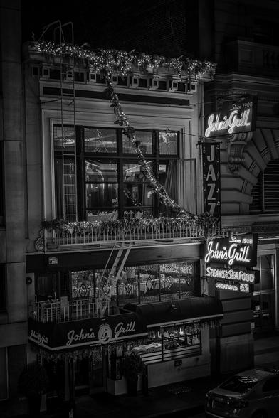 Front of John's Grill by night. Light spilling out through the glass windows of the restaurant is joined by the glow of two neon signs. If you look closely, you can see a photograph of Humphrey Bogart holding the falcon on the wall.