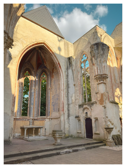 Interior view of a church ruin with tall Gothic arches, partially preserved frescoes, and windows without glass. The altar and stone structures remain in place, surrounded by weathered walls with faded decorative patterns. Sunlight highlights the worn stone, while trees are visible through the open windows under a partly cloudy sky.