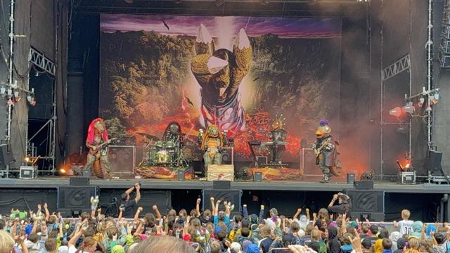 Eine farbenfrohe Live-Performance mit Musikern in einzigartigen Tierkostümen auf der Bühne, mit einem dramatischen Hintergrundbild einer großen Kreatur. Die Menge ist engagiert und hebt aufgeregt die Hände.