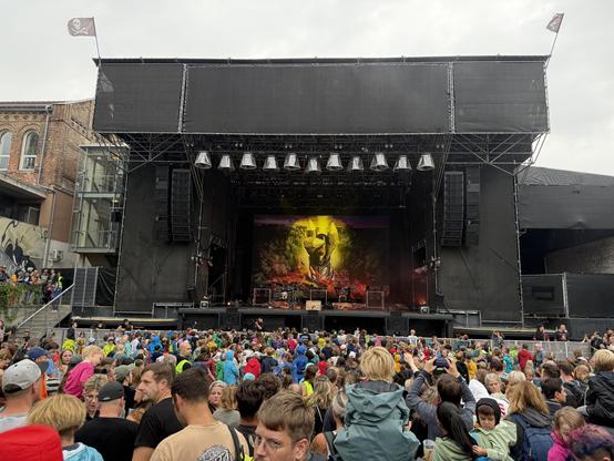 Eine große Menschenmenge versammelt sich vor einer Konzertbühne der Arena Wien mit einer farbenfrohen Kulisse. Das Publikum trägt verschiedene Regenmäntel und genießt die Veranstaltung. Flaggen sind oben auf der Bühne zu sehen, und die Atmosphäre ist trotz bewölktem Wetter lebendig.