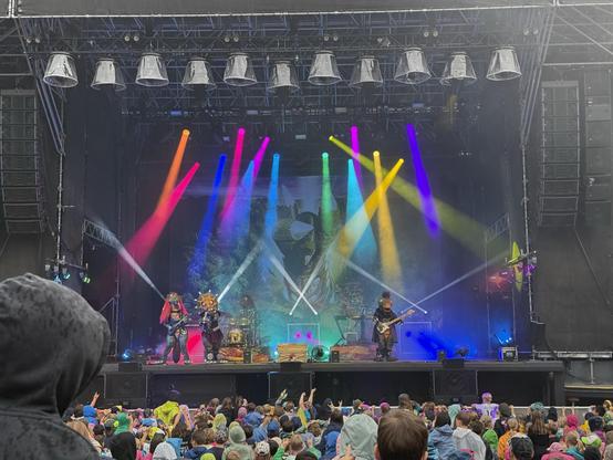 A colorful concert scene featuring performers in elaborate costumes on stage, with vibrant stage lighting. A crowd of spectators, many in raincoats, is visible in front of the stage.