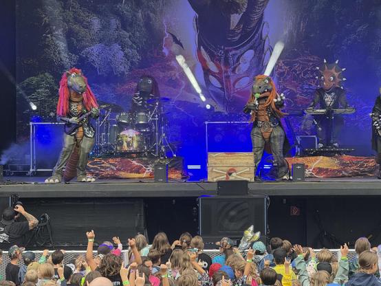 Eine lebhafte Aufführung mit Musikern in bunten Dinosaurierkostümen auf der Bühne, mit einem lebendigen Hintergrund, der einen Wald und eine Vulkanlandschaft darstellt. Das Publikum unten engagiert sich begeistert von der Aufführung.