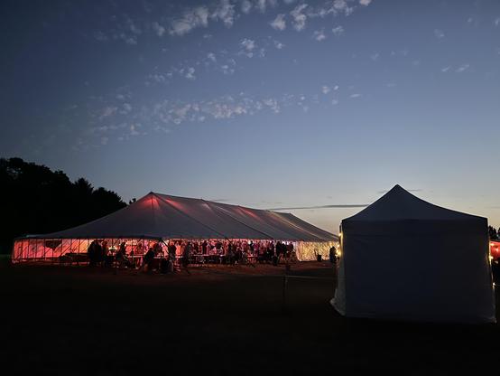 A large marquee lit with coloured lights, at dusk under a beautiful evening sky. People are dancing and listening to a band