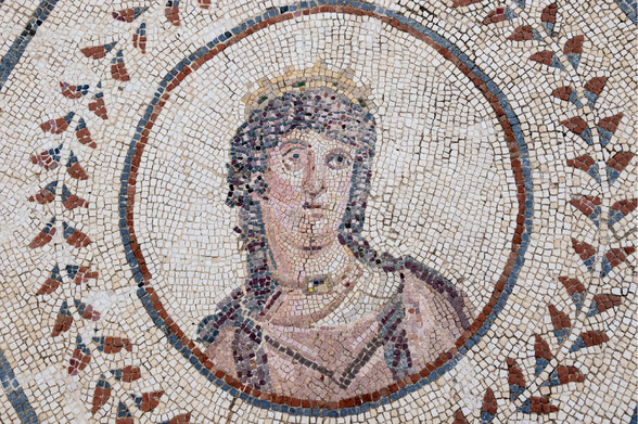Mosaic of the bust of Venus inside a circle. She wears a golden crown, her dark hair flowing over her shoulders. She wears a golden necklace with a rectangular gemstone in the middle.