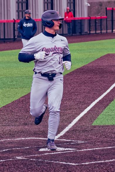 Baseball player wearing grey clothesthat says Quakers 16 puts his foot on home plate on a brown stripe in the field surrounded by green, behind him a man wears jblue jacket that says Quakers is looking on.