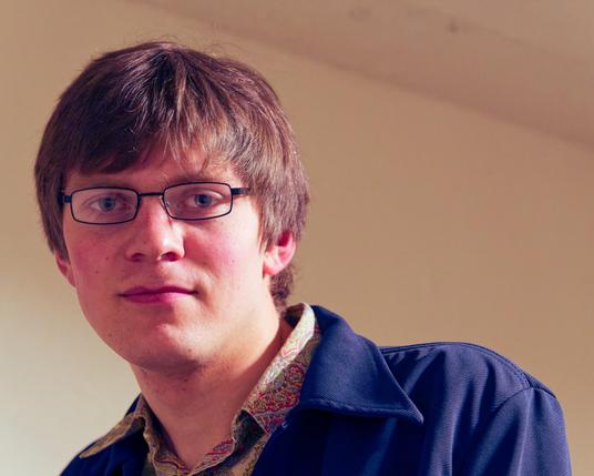 Young man with short hair,  thin glasses,  and a paisley color shirt mostly covered by a blue jacket.