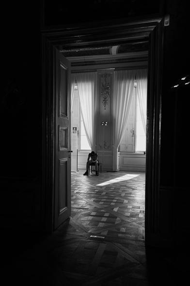 Scene from Nymphenburg Palace in Munich. View through a door on a boy sitting on a chair, looking at a smartphone. Light is falling on the florr from two big windows behind him.