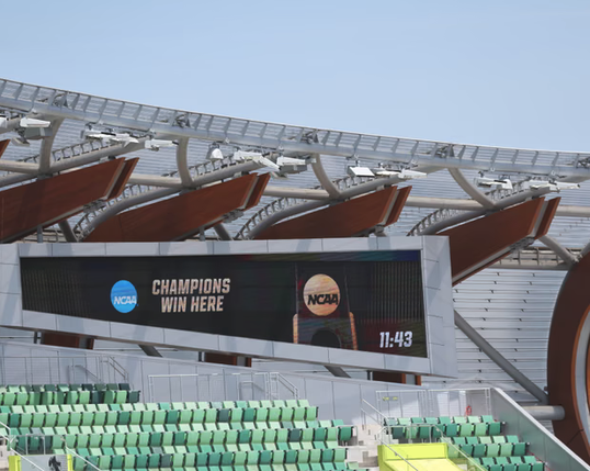Empty stadium with green seats below and some big curved brown things above and a trapezoidal digital sign that says (NCAA) CHAMPIONS WIN HERE and has the time 11:43