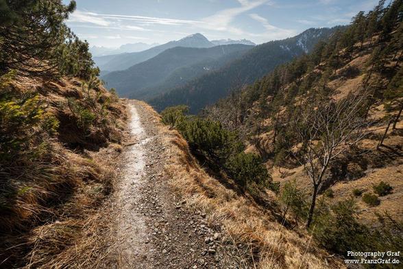 A scenic dirt path winds its way through a rugged mountain landscape, surrounded by lush greenery and rocky terrain. The path is flanked by tall trees and bushes, with a backdrop of towering mountains and a cloudy sky. The earthy tones of brown and black dominate the scene, creating a natural and serene atmosphere. This outdoor mountain trail offers a peaceful and tranquil setting, perfect for hiking and exploring the wilderness. The image captures the essence of the highland wilderness, showca…