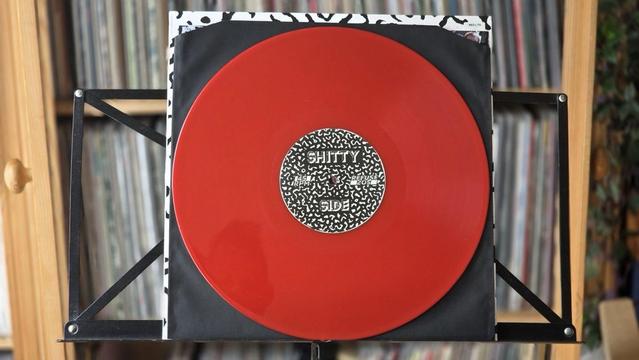 Eine rote Schallplatte wird auf einem schwarzen Ständer präsentiert, auf dem ein Etikett mit der Aufschrift "SHITTY SIDE" prangt. Die Platte befindet sich in einer Hülle mit einem abstrakten schwarz-weißen Muster, während im Hintergrund Regale mit weiteren Platten zu sehen sind.