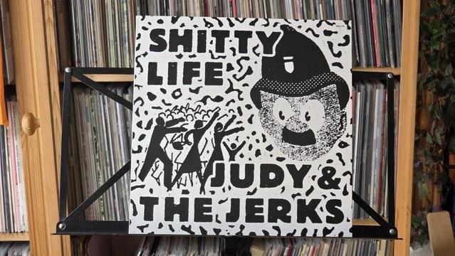 Das Bild zeigt ein Plattencover mit dem Titel "SHITTY LIFE" von "JUDY & THE JERKS". Das Design umfasst fetten schwarzen Text, Illustrationen von Figuren und eine Cartoon-Figur mit Schnurrbart und Hut.