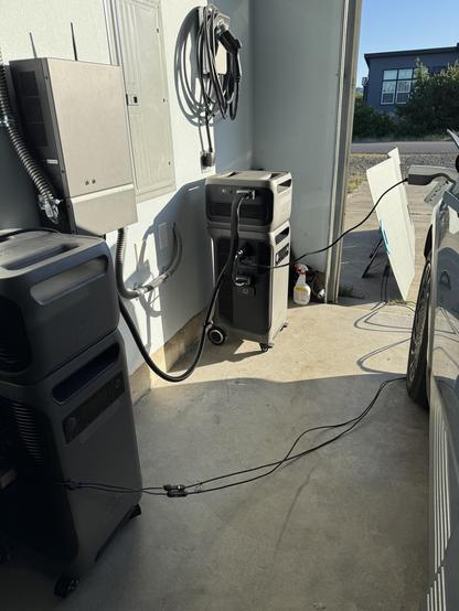 A garage setup featuring two Anker Solix batteries with expansion batteries atop them, connected to a home Power Panel, which is connected to an electrical subpanel. One power station is being charged by a solar panel outside the garage on the concrete driveway. Thin black cables run along the floor from one battery to the PV panel. A standard 120V AC power cord connects the other battery to a V2L dongle on a blue-gray EV in the garage. An EV charger with cable looped around it is mounted on the wall. 