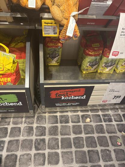 Säcke mit Grillkohle, die in einem Regal ausgestellt sind, auf dem normalerweise Kartoffeln liegen. Das Regal ist mit „vorwiegend festkochend“ bezeichnet