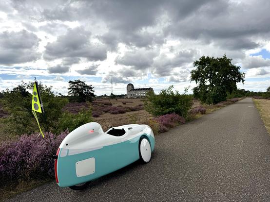Een wit-blauwe velomobiel DF met groen vlaggetje staat op een asfaltweg met links er naast paars bloeiende heide. Wat verder weg staan twee struiken en een boom en daar tussendoor zie je het zendergebouw van Radio Kootwijk vanaf de linker achter-zijkant. De lucht is deels bedekt met donkergrijze wolken, maar er is ook wat blauw. 