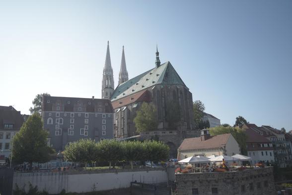 Ein horizontal aufgenommenes Foto einer Stadtansicht an einem hellen Tag. Im Zentrum des Bildes steht eine große, gotische Kirche mit einem markanten grünen Dach und zwei schlanken, hohen Kirchtürmen mit spitzen Aufsätzen. Rechts von der Kirche sind Dächer von Wohnhäusern zu sehen. Im Vordergrund befindet sich eine befestigte Terrasse mit Bäumen, auf der mehrere runde Tische mit weißen Sonnenschirmen stehen, die zu einem Restaurant gehören. Der Himmel ist klar und hellblau.