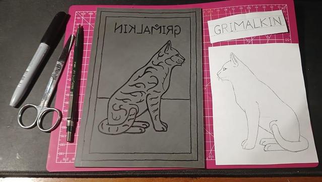 Preparation of a cat lino print. So far the design of the cat has been transferred onto the lino on the left and the outline has been gone over with a permanent marker. On the right is the pencil drawing of the cat that was transferred to the left and reversed. There is text "Grimalkin" reversed on the left and the right way round in pencil on a separate piece of paper on the right. There is also the permanent marker, a pair of scissors, and a mechanical pencil all the way to the left of the picture.