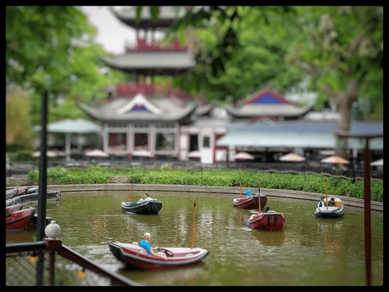 Several people in boats, in a lake. The tilt shift effect makes them look like toys or models. 
