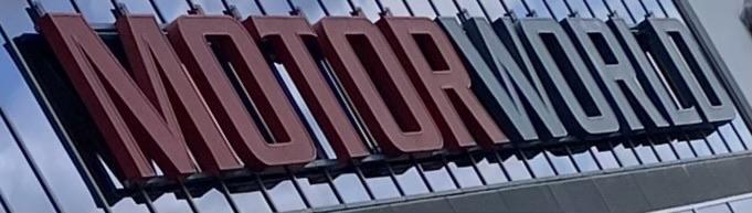 The image features a close-up of a sign that reads "MOTOR WORLD," with bold, modern lettering in red and black against a reflective surface.