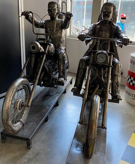 Two large, metal sculptures of bikers sitting on motorcycles. The figures are intricately designed with mechanical parts, and they each wear distinctive outfits. One has a leather jacket, while the other features a long coat. The motorcycles are displayed on a stand
