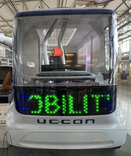 An innovative autonomous vehicle featuring a transparent design and a bright LED display showing the word "MOBILITY." The interior includes a modern red seat. The vehicle is displayed in a spacious exhibition area.